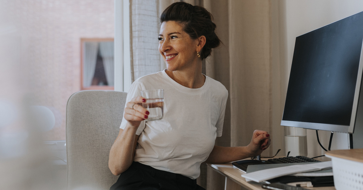 En leende kvinna vid ett skrivbord med dator och blanketter håller i ett vattenglas och vänder sig mot rummet. 