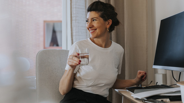 En leende kvinna vid ett skrivbord med dator och blanketter håller i ett vattenglas och vänder sig mot rummet. 