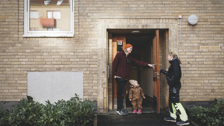 En ung pappa på väg ut från en port tillsammans med sin lilla dotter håller upp dörren för en fastighetsarbetare på Mölndalsbostäder.