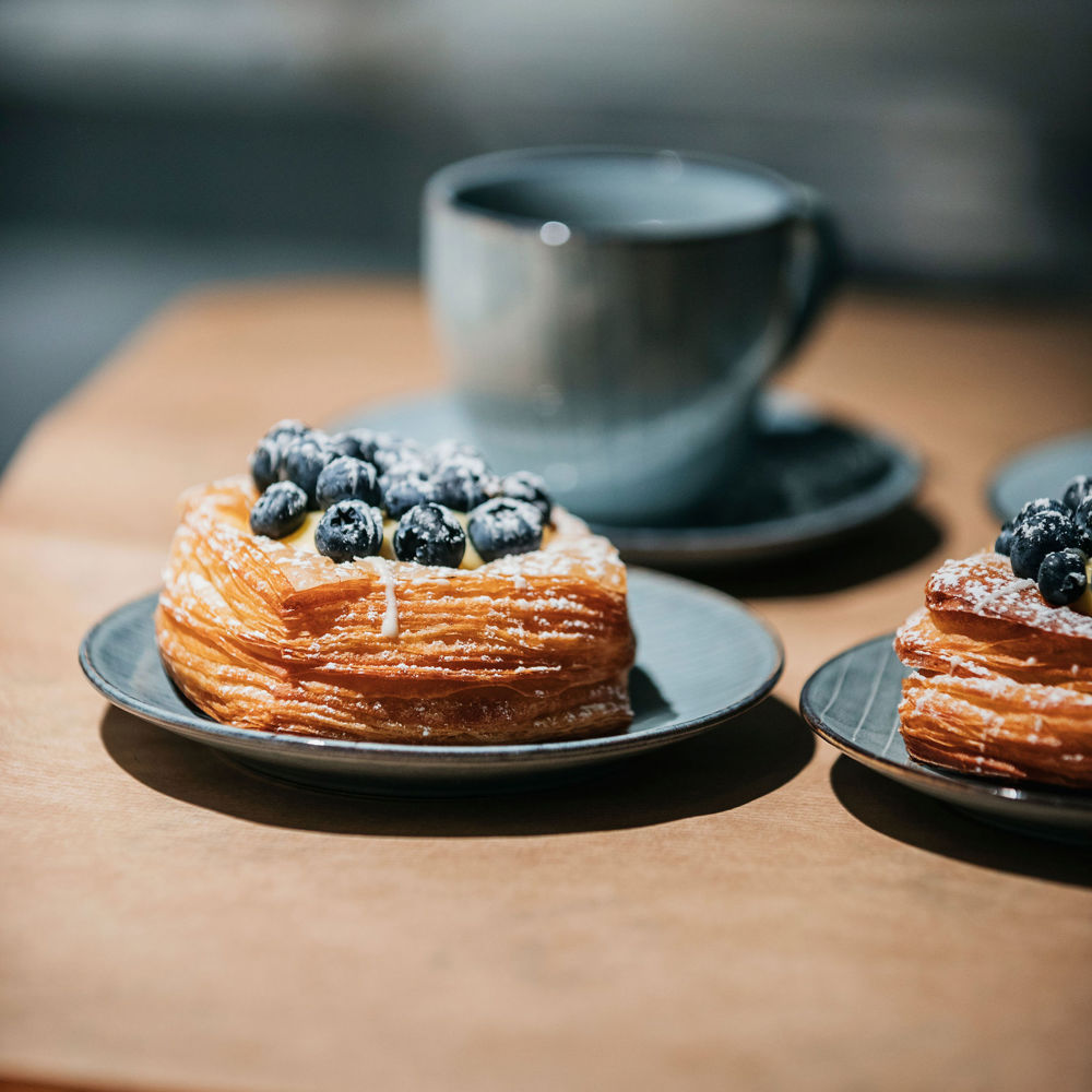 Närbild på en blå kaffekopp och ett fat med en bakelse toppad med färska blåbär. 