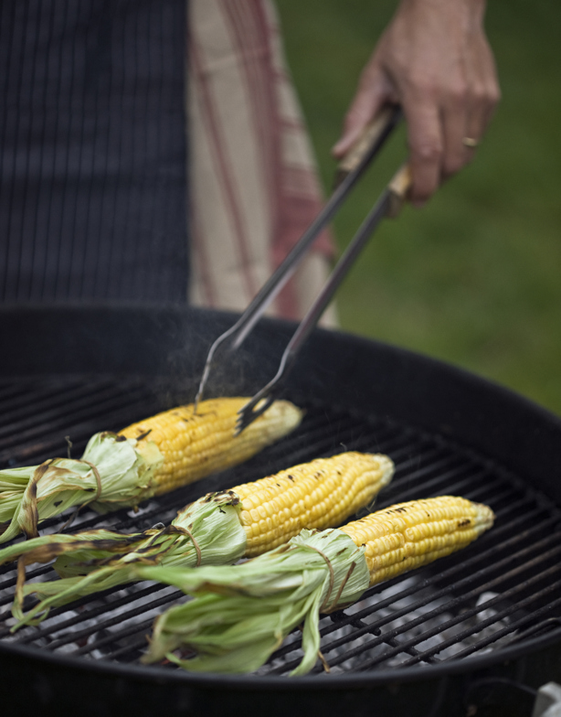 en grill i förgrunden med tre grillade majskolvar. En hand håller i en tång.