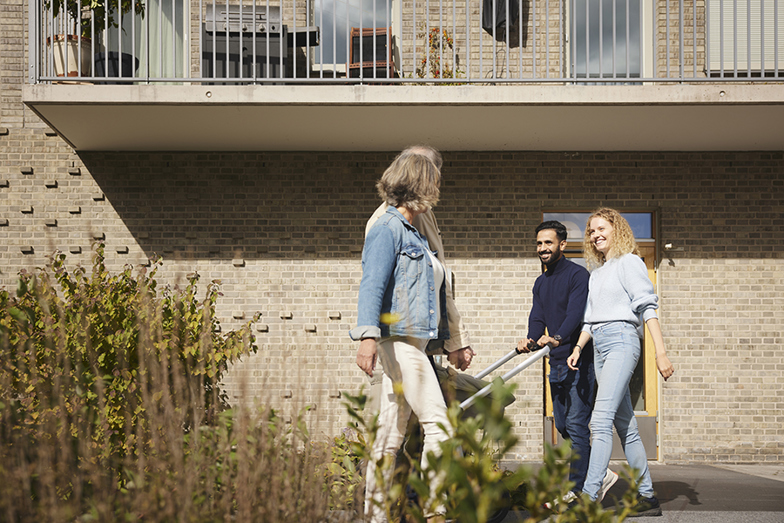 Ett äldre par med en kvinna och man möter ett yngre par med kvinna, man och en barnvagn utanför ett hyreshus. De yngre paret ler mot de äldre.