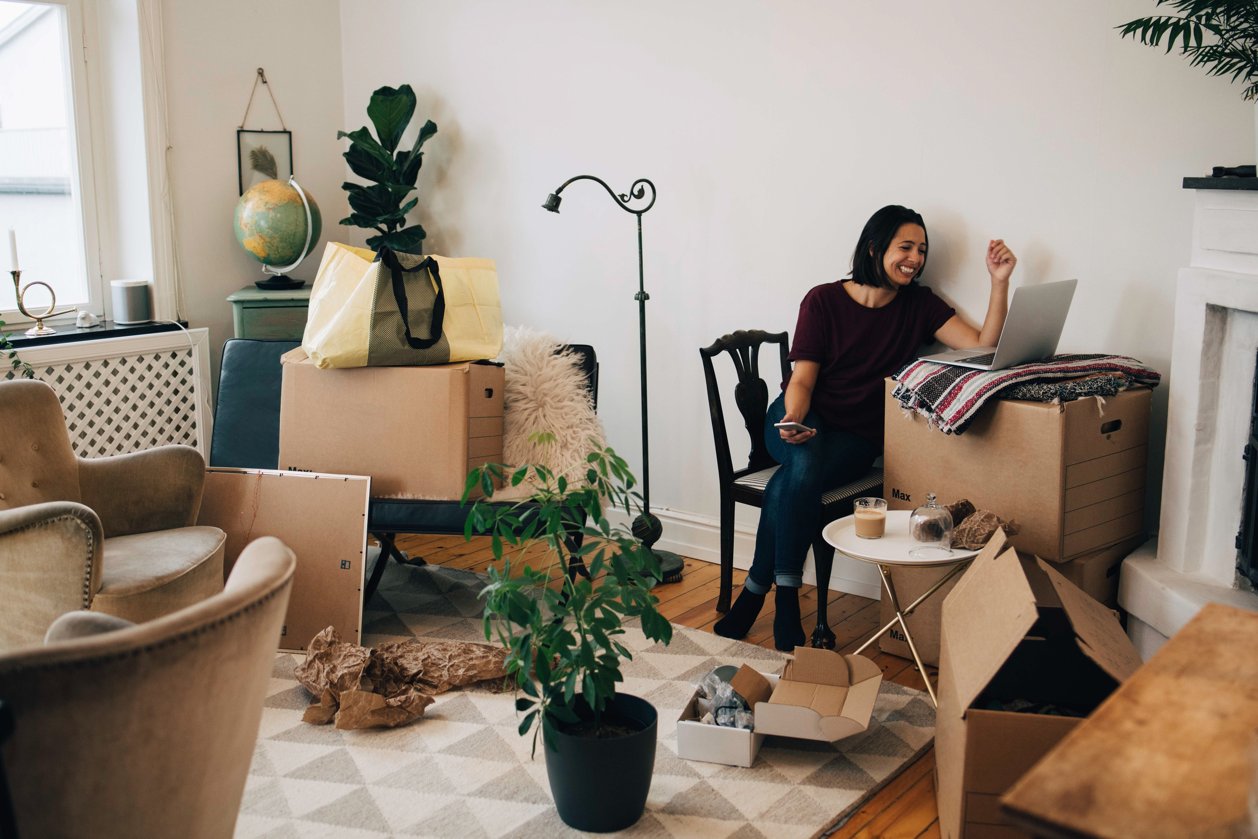 Ung leende kvinna sitter vid en laptop som är placerad på en av flyttkartongerna i vardagsrummet. 