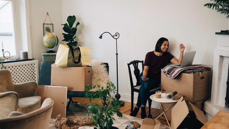 Ung leende kvinna sitter vid en laptop som är placerad på en av flyttkartongerna i vardagsrummet. 