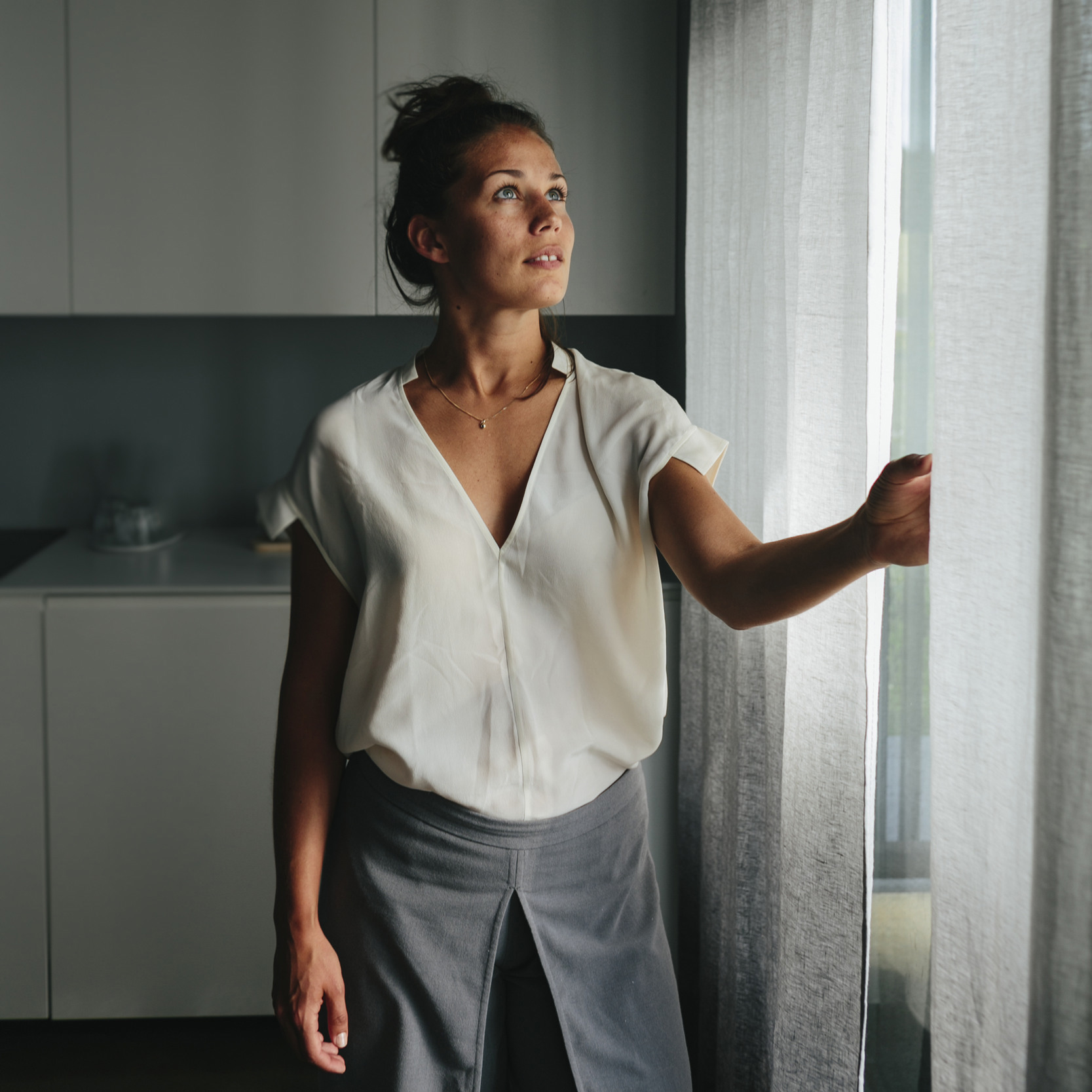 En ung mörkhårig kvinna står vid ett fönster i ett vitt hem och drar för en gardin. 