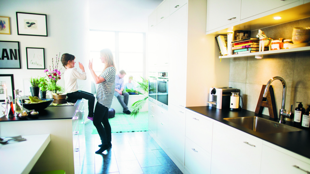 En mamma leker med sitt barn som sitter på en köksbänk i ett vitt öppet kök med en pappa och ett barn lite suddigt i bakgrunden. 