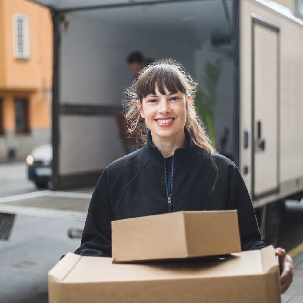 Leende ung kvinna bär flyttlådor från en mindre lastbil i bakgrunden