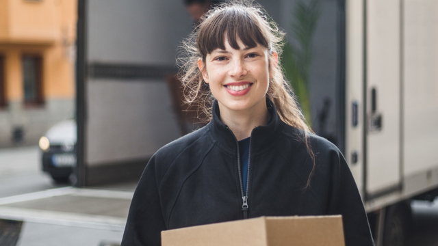 Leende ung kvinna bär flyttlådor från en mindre lastbil i bakgrunden
