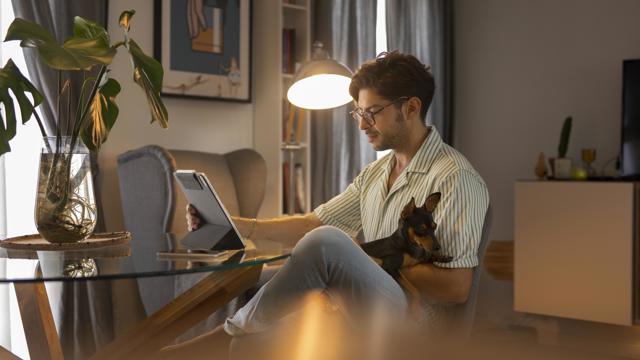 En man sitter med sin hund i knät vid ett bord och tittar på sin Ipad i skenet av en lampa. 