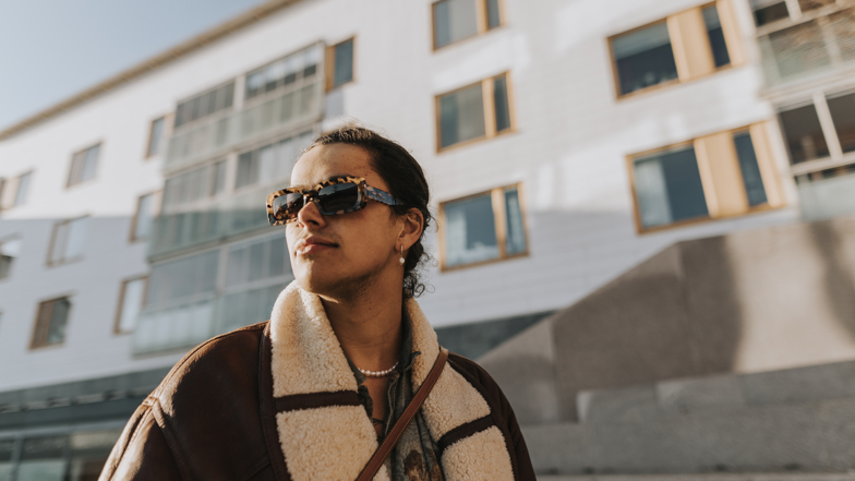 En ung person i solglasögon och brun skinnjacka går framför ett flerfamiljshus. 