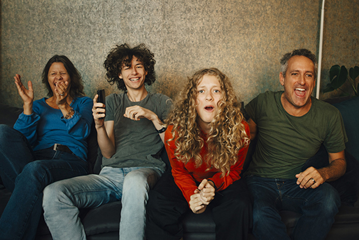 En glad familj med mamma, två ungdomar och en pappa tittar på tv tillsammans i en soffa.